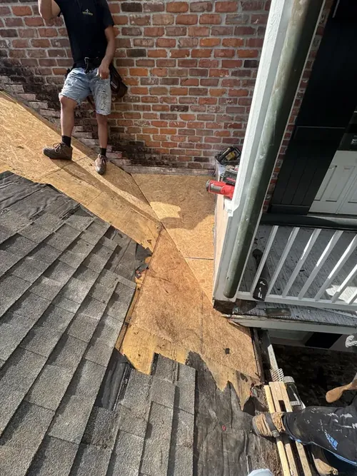 A Cypress Roofing crew member laying fresh decking at a roof edge against a brick chimney wall